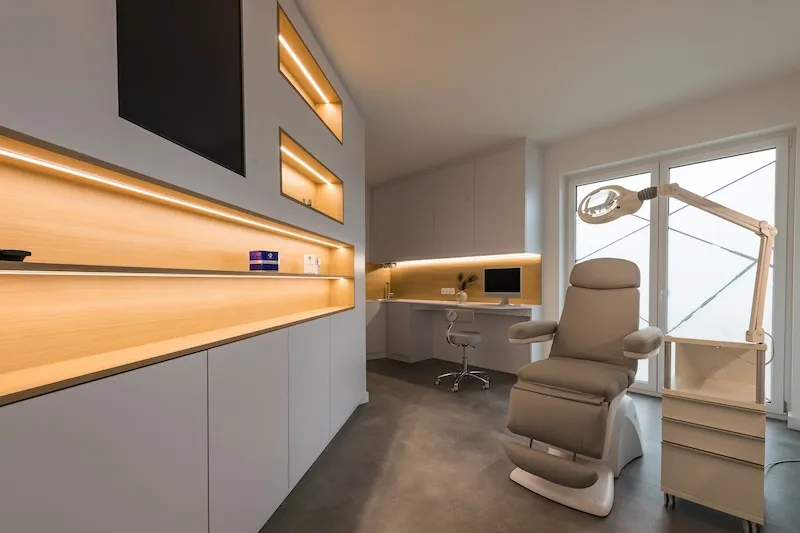 Modernes Behandlungszimmer des Praxisstandort Landsberg am Lech mit einem grauen Behandlungsstuhl, beleuchteten Regalen und einem Bildschirm auf einem weißen Schreibtisch.