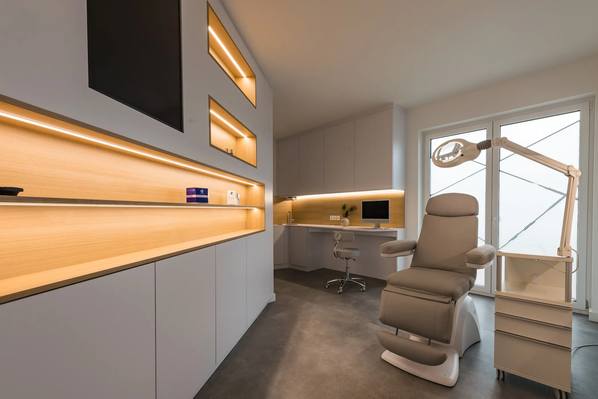 Modernes Behandlungszimmer des Praxisstandort Landsberg am Lech mit einem grauen Behandlungsstuhl, beleuchteten Regalen und einem Bildschirm auf einem weißen Schreibtisch.