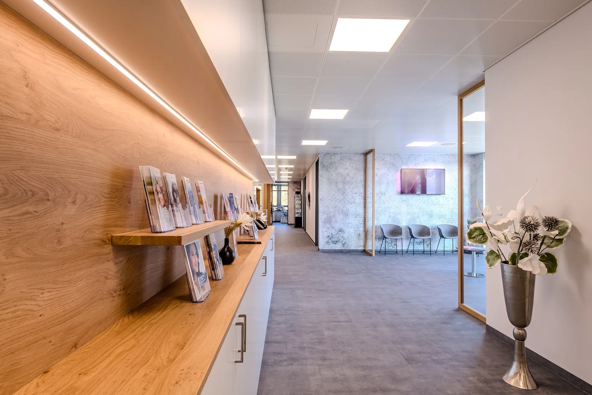 Moderner Wartebereich mit grauen Stühlen, Holzregal mit Broschüren und einer Vase mit künstlichen Blumen im Flur.