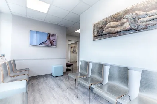 Warteraum mit Holzstühlen entlang der Wände, einem kleinen blauen Sitzwürfel, und einem Fernseher an der Wand, der eine Frau mit einem Kosmetikgerät zeigt.