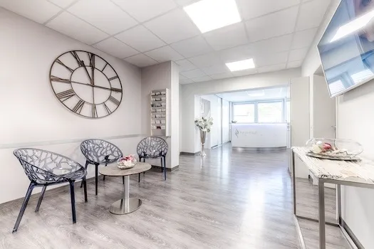 Moderner Wartebereich mit drei schwarzen Stühlen, kleinem runden Tisch mit Dekoration, großer Wand-Uhr mit römischen Zahlen und hellem Boden.