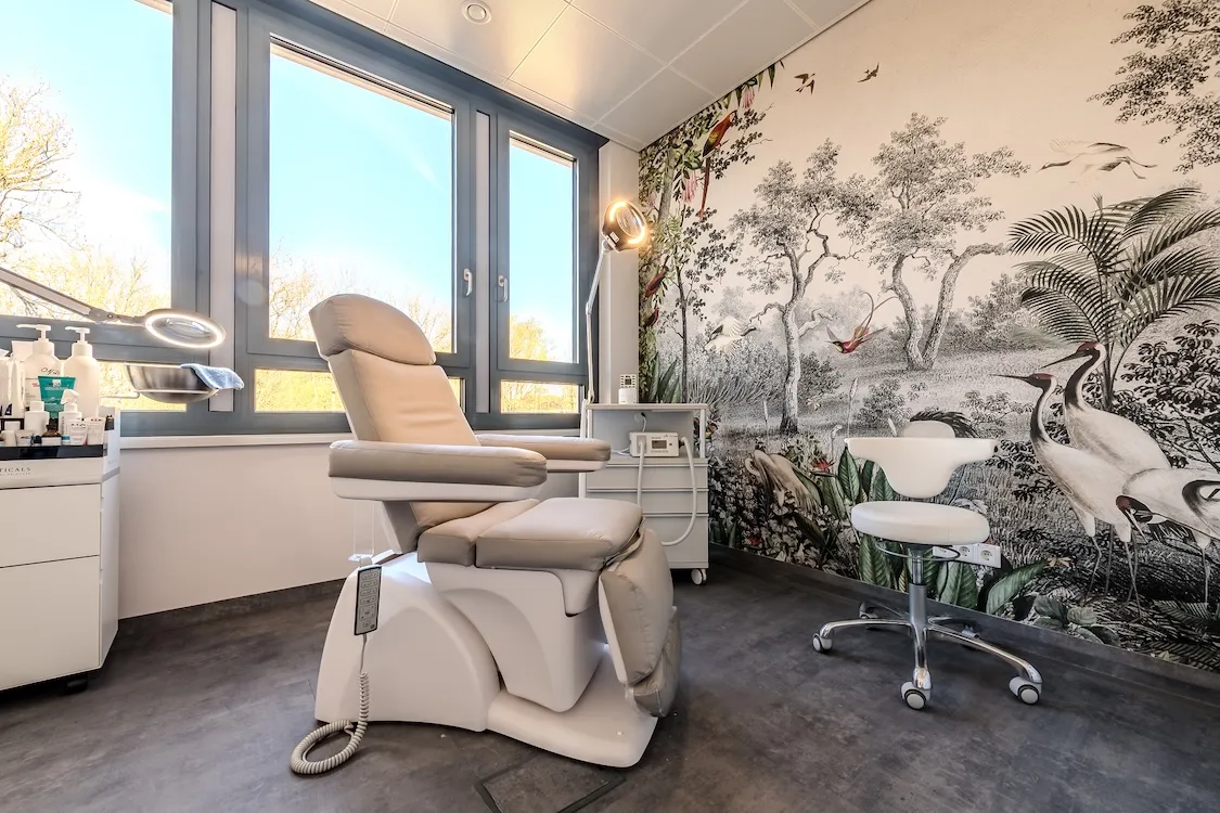 Behandlungszimmer mit einem bequemen weißen Klinikstuhl und weißem Hocker neben einer Wand mit Tier- und Baumzeichnung.