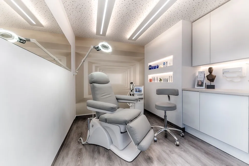 Moderner Behandlungsraum mit grauem Behandlungsstuhl, Klapprollhocker, Wandregalen mit Kosmetikartikeln und dekorativer Wandgestaltung.