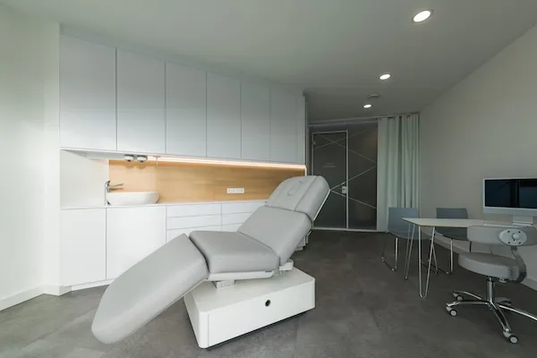 Moderner Behandlungsraum mit grauem verstellbarem Untersuchungsliege, weißen Schränken und Schreibtisch mit zwei Stühlen.
