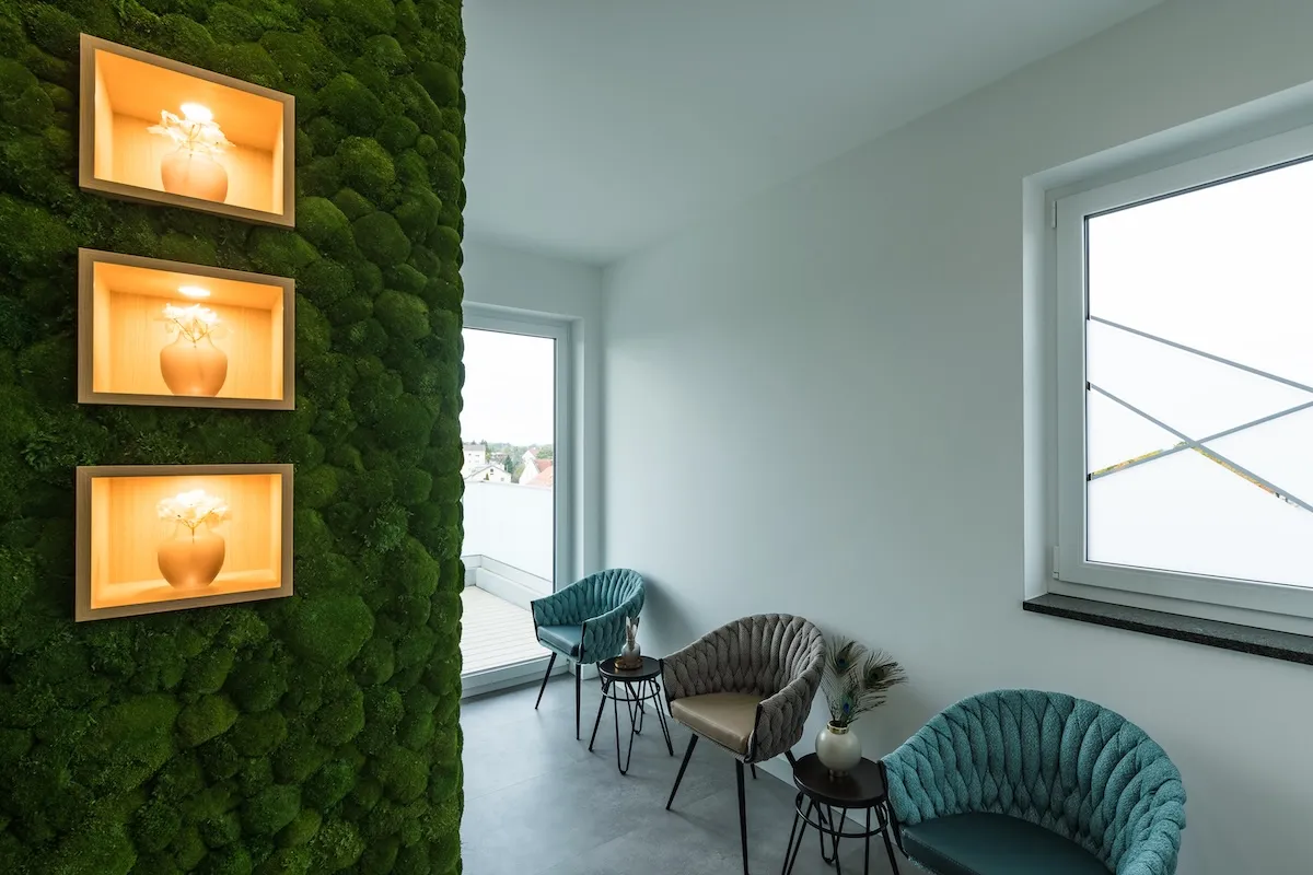 Wartebereich mit drei gepolsterten Stühlen, kleinen Tischen und grüner Mooswand mit beleuchteten Nischen, helle Wände und Fenster.