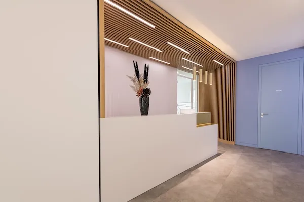 Moderner Empfangsbereich mit weißem Empfangstresen, einer Vase mit Trockenblumen und einer Holzwanddecke mit eingebauten Leuchten.