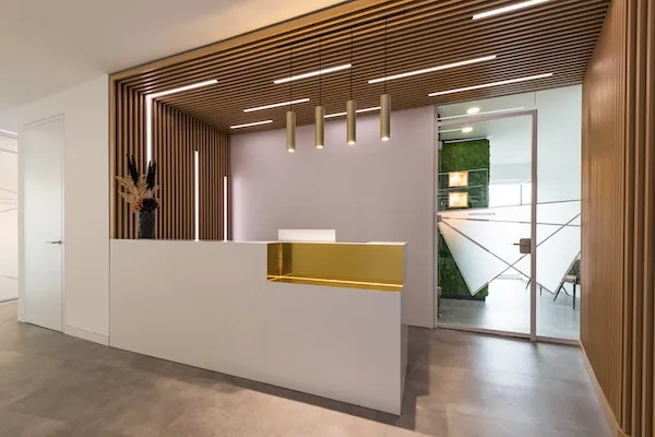 Moderner Empfangsbereich mit weißem Tresen, goldener Akzentleiste, Holzverkleidung an Decke und Wänden sowie einer Glastür im Hintergrund.
