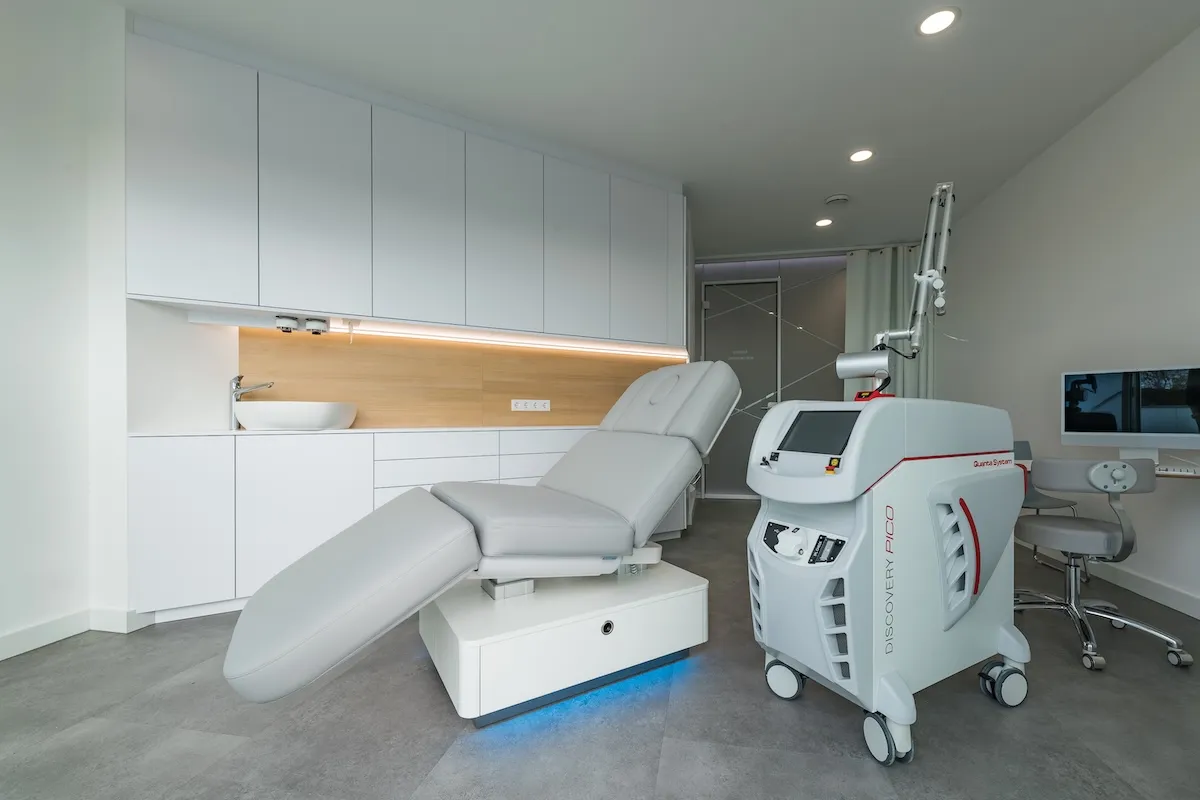 Moderne Behandlungszimmer mit grauem Liegestuhl, Lasermaschine Discovery Pico und weißen Schränken.