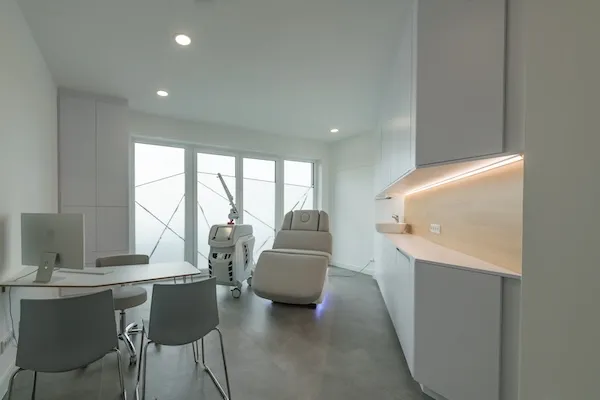 Moderne, helle Arztpraxis mit grauen Stühlen, einem weißen Schreibtisch, einem Behandlungsstuhl und medizinischem Gerät vor frosted Fenstern.