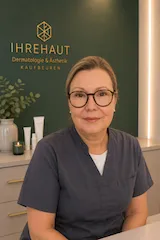 Porträt einer Frau mit Brille in grauer medizinischer Arbeitskleidung vor IHREHAUT Praxisschild.