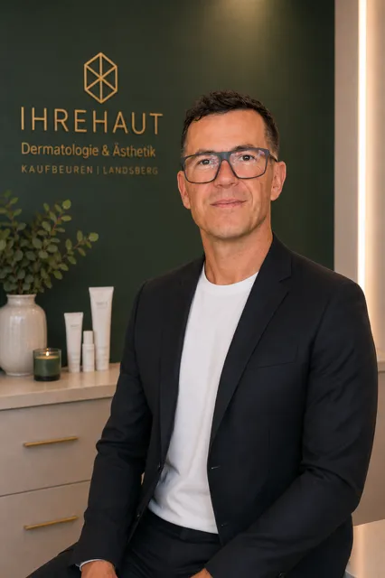 Mann mit Brille und Anzug vor Wand mit Logo von Hautklinik für Dermatologie und Ästhetik.
