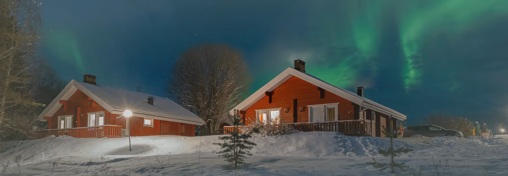Two red wooden cabins in snowy landscape under a night sky with visible green northern lights.