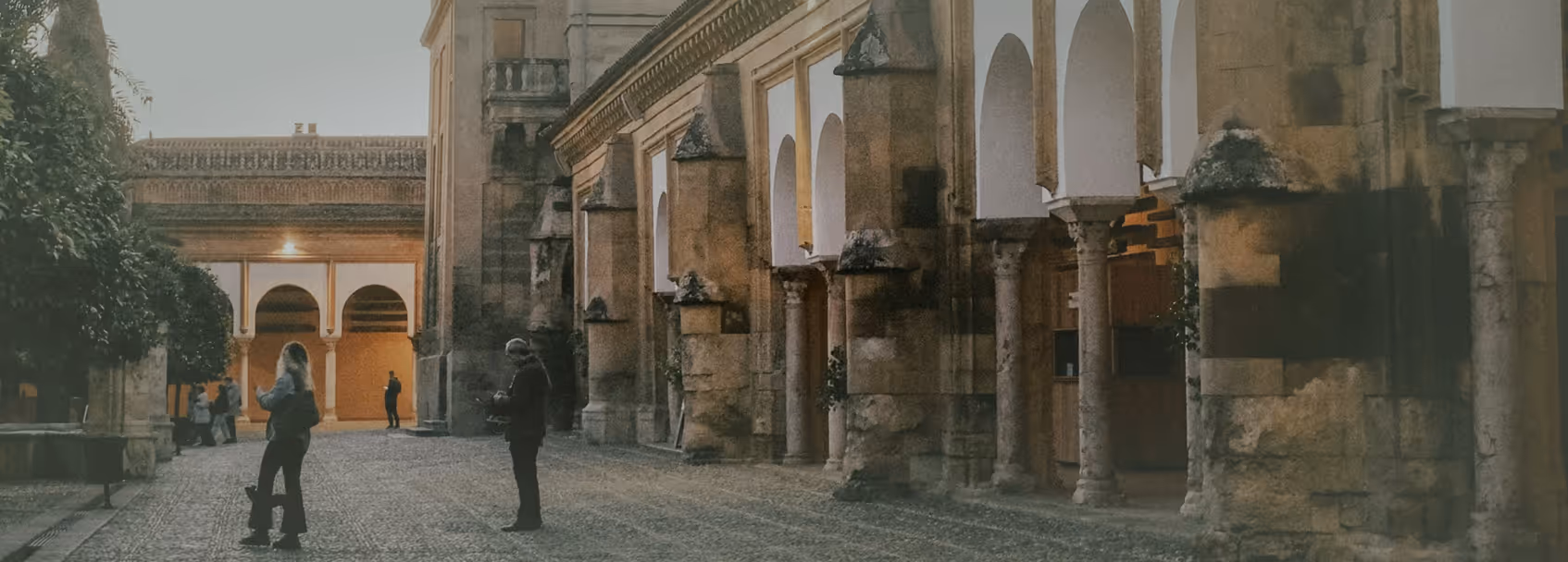 People standing and taking photos in a historic courtyard with stone arches and columns during dusk.