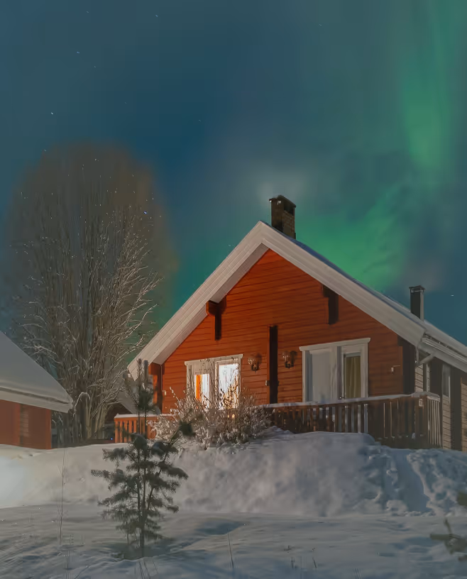 Red wooden house with snow-covered yard and northern lights glowing in the night sky.
