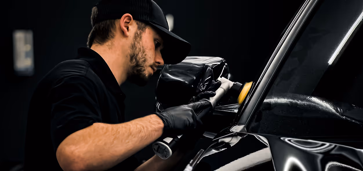 Mand med sorte handsker og kasket polerer en sort bildør med en håndholdt roterende polermaskine