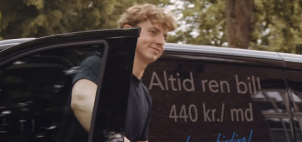 Ung mand i sort t-shirt åbner døren på en sort bil med dansk tekst om priser på siden