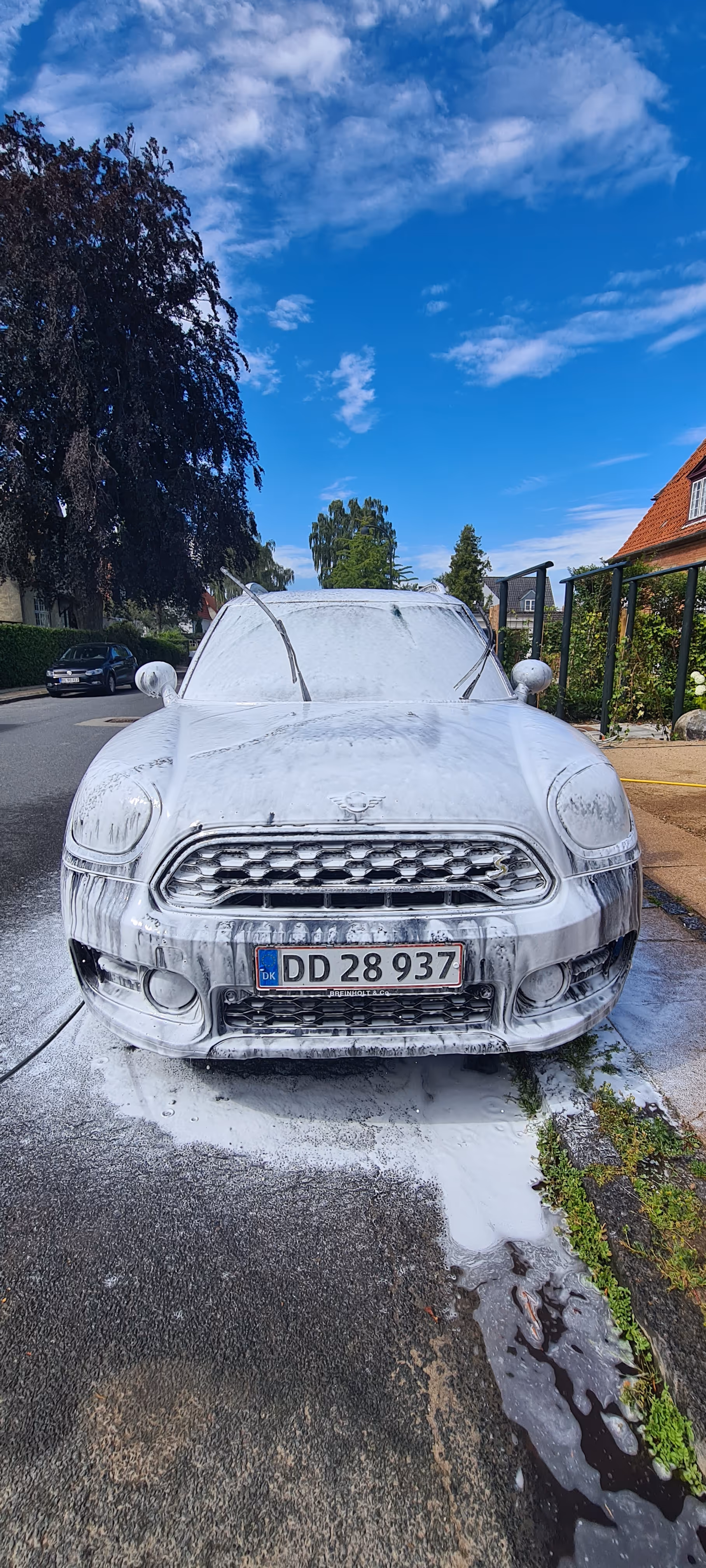 Mini Cooper set forfra dækket af hvidt bilvaskeskum på gade med blå himmel og træer i baggrunden