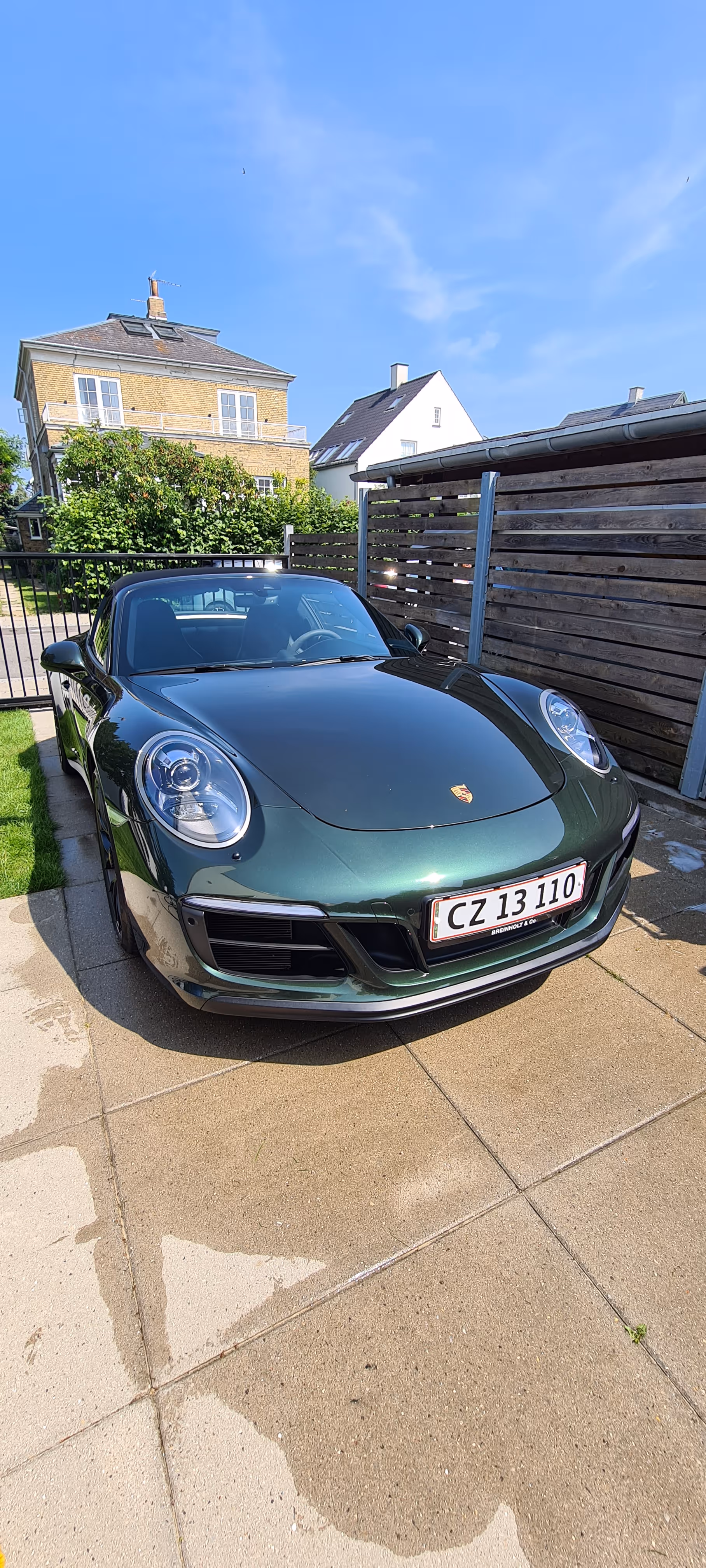 Mørkegrøn Porsche sportsvogn parkeret på en indkørsel med bolighuse og en klar blå himmel i baggrunden