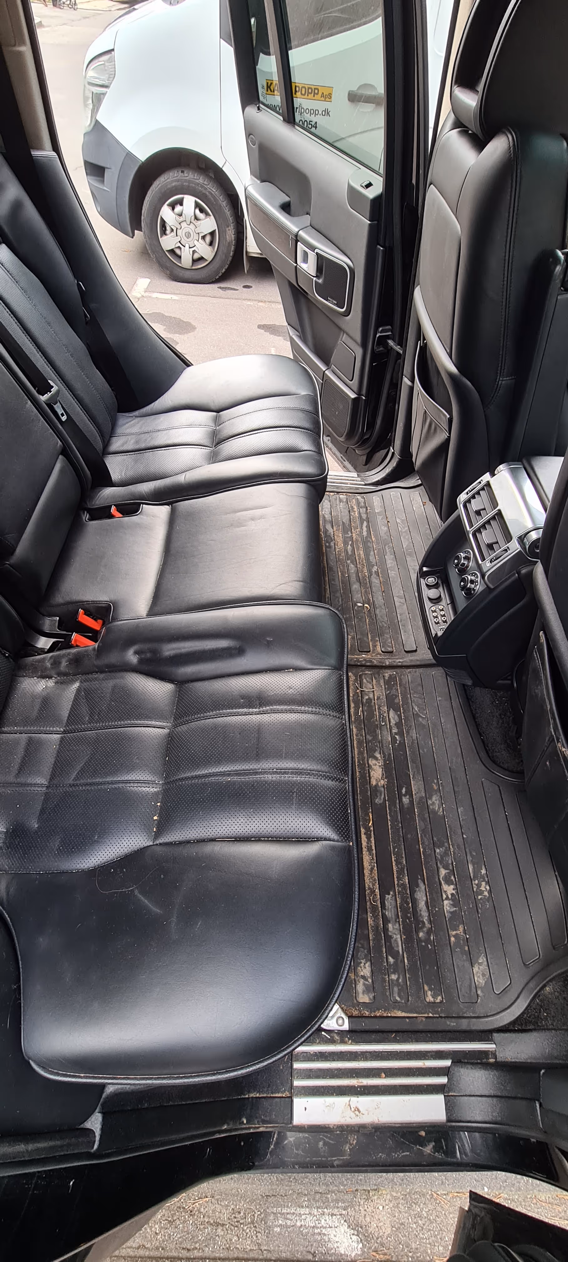 Sort lædersæde på bagsædet i en bil med døren åben, der viser et beskidt gummi-gulvtæppe og en del af en hvid varevogn udenfor
