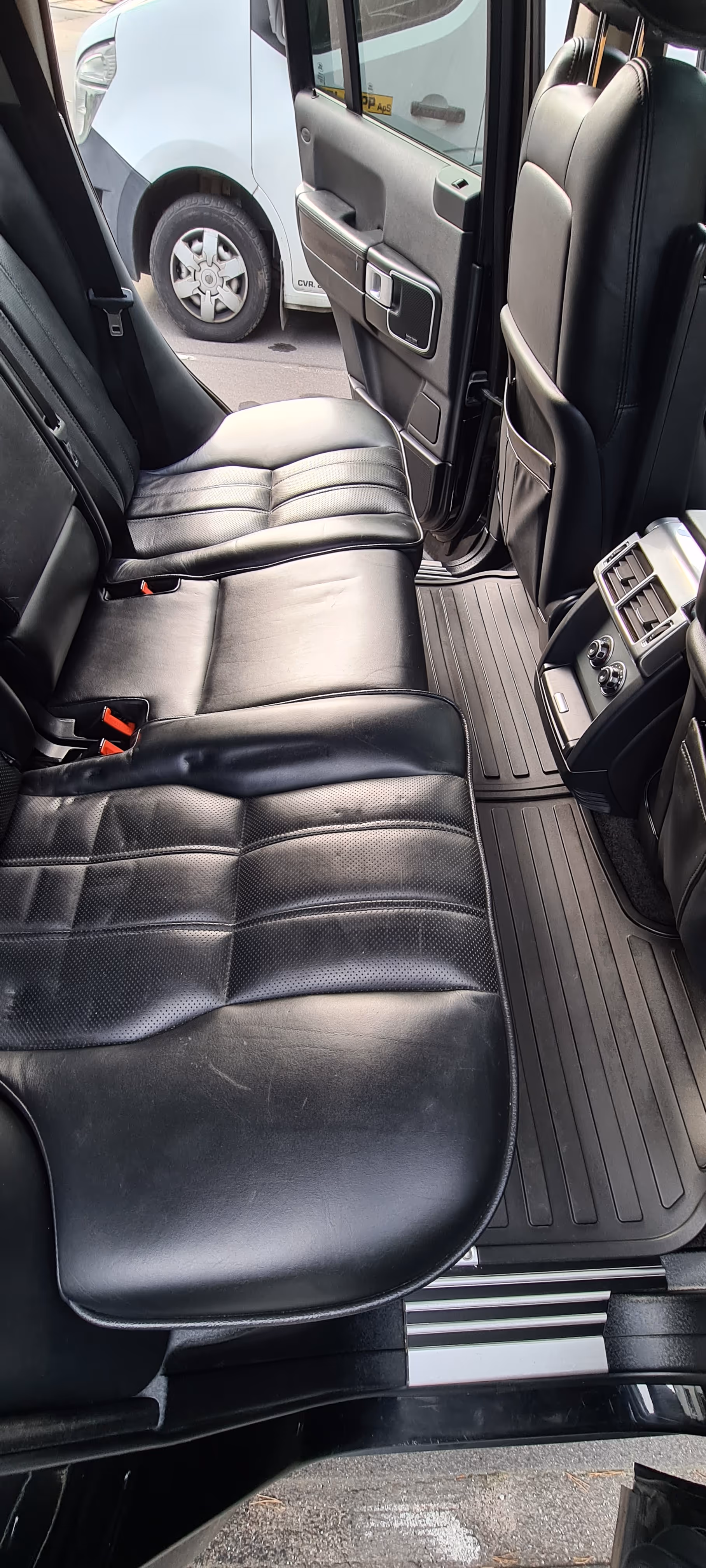 Sort lædersæde på bagsædet i en bil med døren åben, der viser et gummi-gulvtæppe og en del af en hvid varevogn udenfor