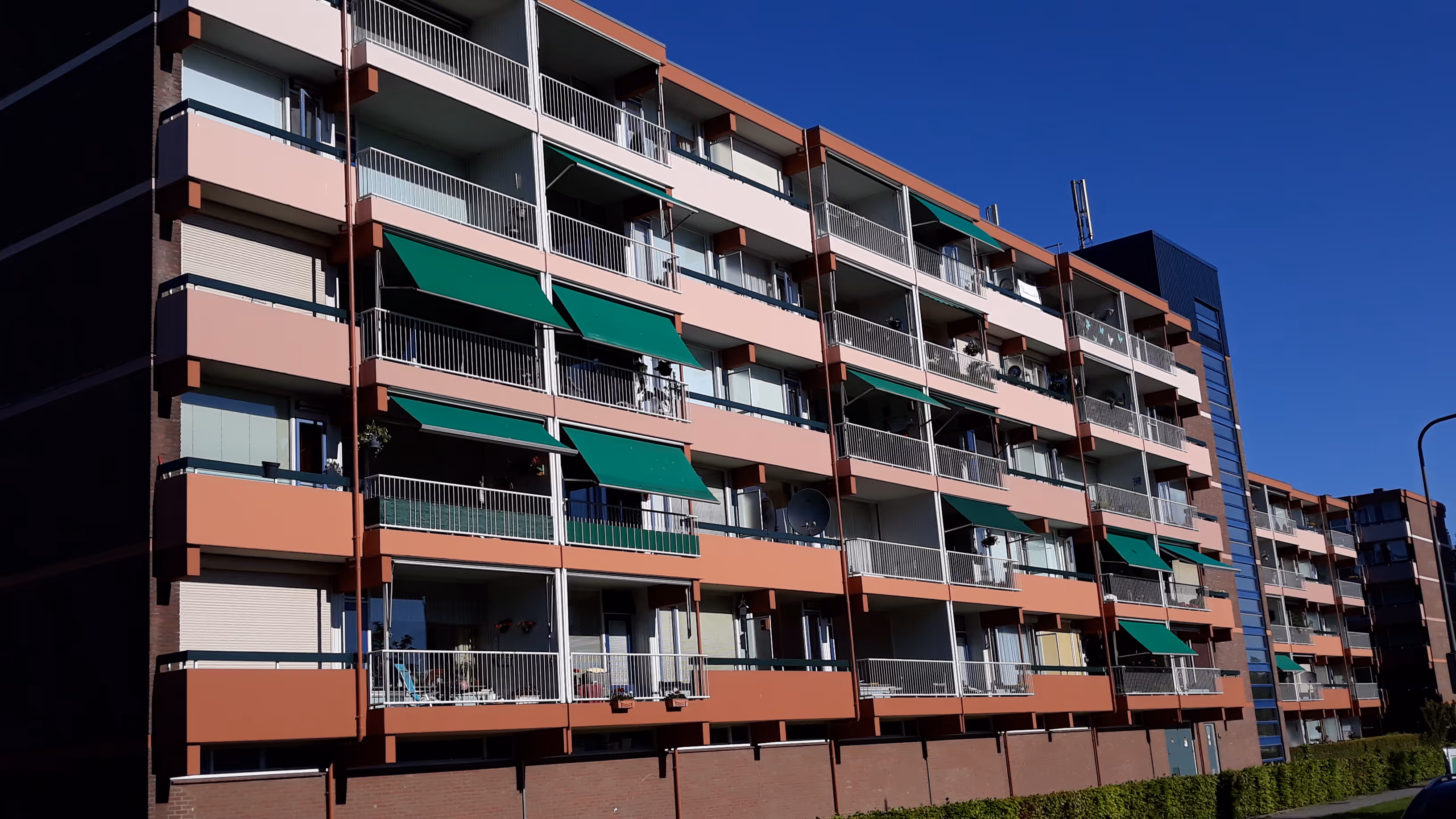 Modern appartementencomplex met oranje en witte gevels en groene zonneschermen onder een heldere blauwe hemel.