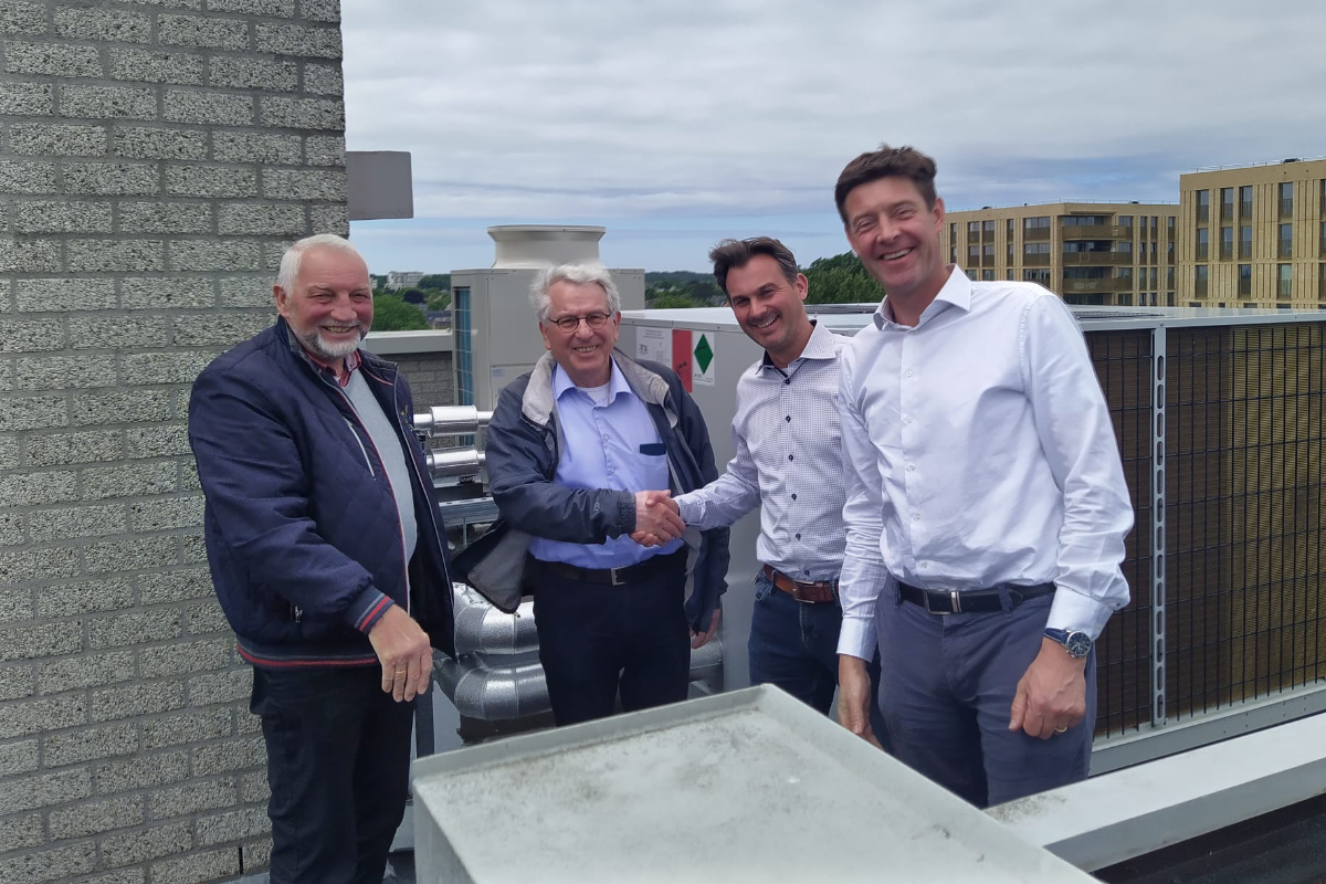 Vier mannen staan buiten op een dak bij een gebouw, twee van hen schudden elkaar de hand terwijl ze glimlachen.