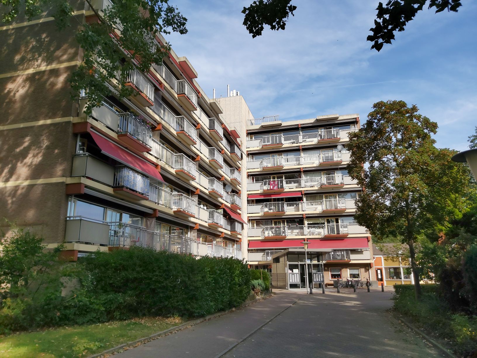 Wooncomplex met balkons en rode zonneschermen onder een blauwe lucht, met bomen en een voetpad ervoor.