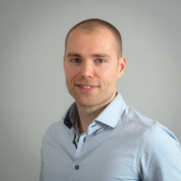 Portret van een glimlachende man met kort haar en een lichtblauw overhemd tegen een grijze achtergrond.