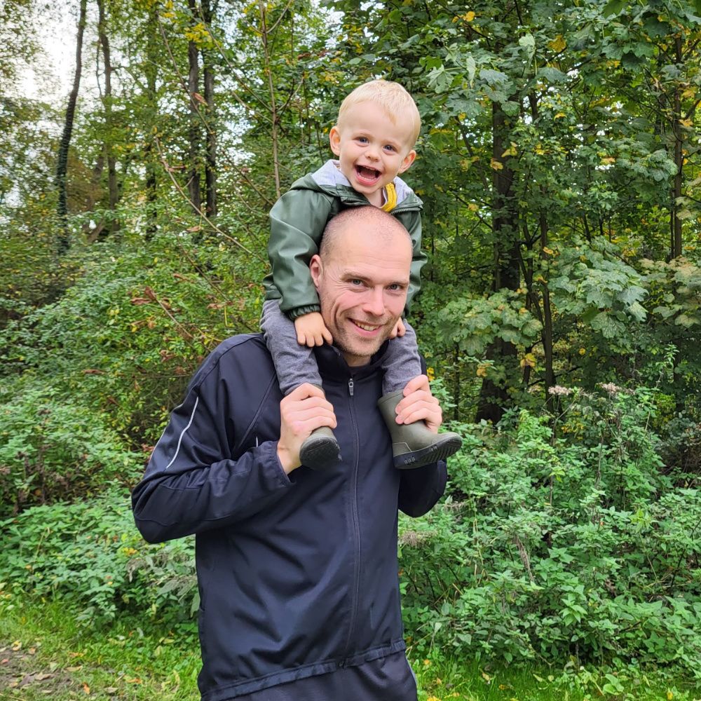 Vader draagt zijn lachende jonge zoon op zijn schouders in een groene bosrijke omgeving.