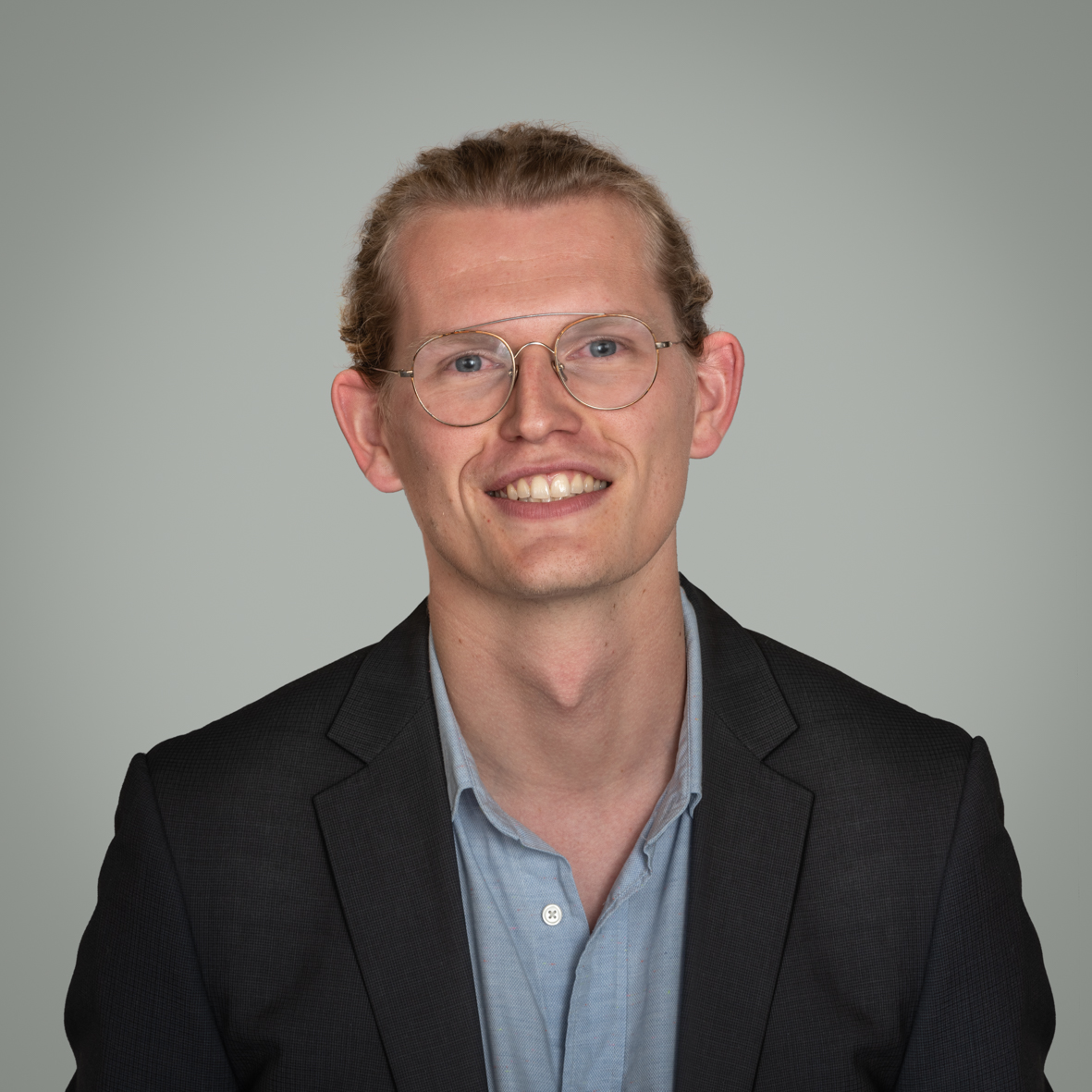 Man met blond haar, een bril en een donker colbertjas lacht naar de camera voor een grijze achtergrond.