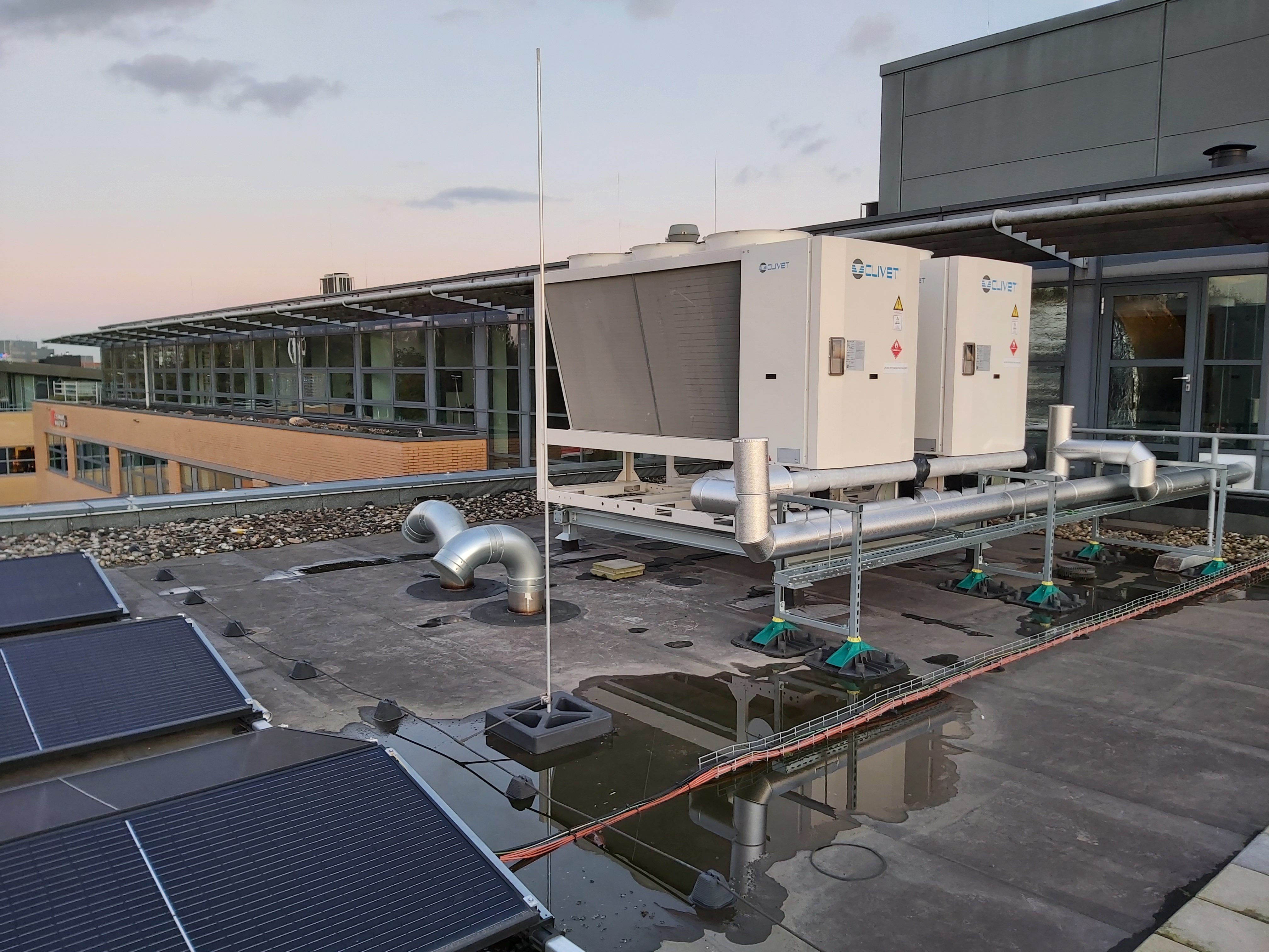 Dak met zonnecellen, waterplassen en twee grote Clivet airconditioningunits bij een modern kantoorgebouw.