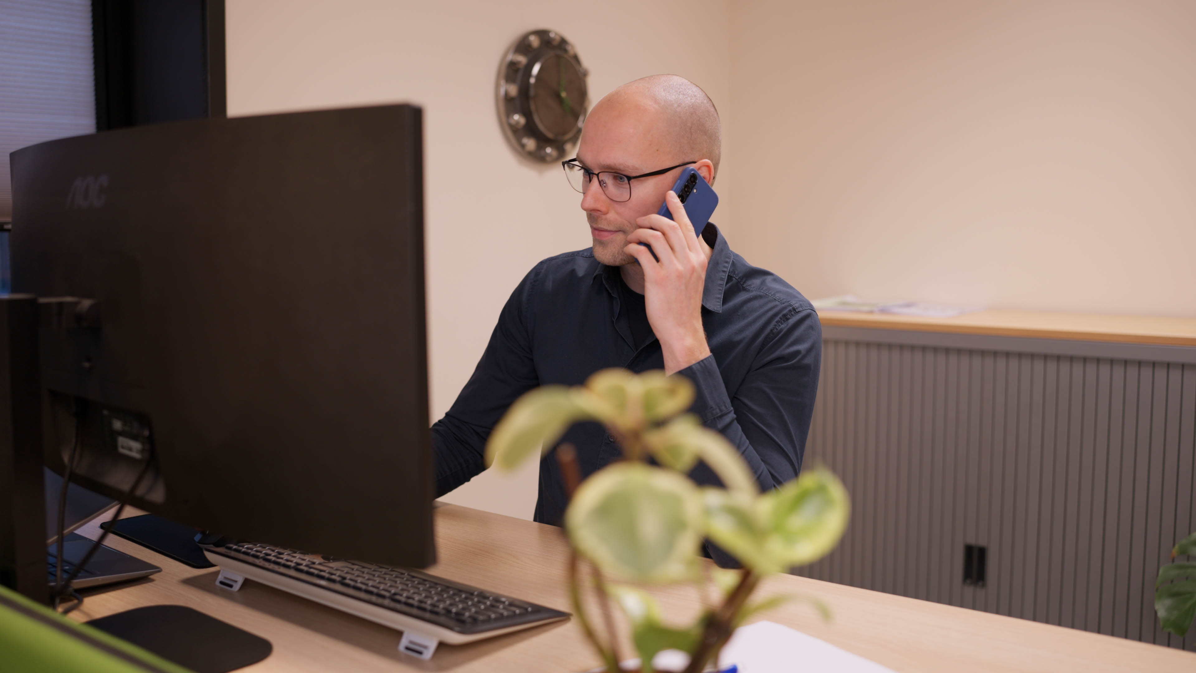 Man met bril aan een bureau die op zijn computer kijkt terwijl hij telefoneert.