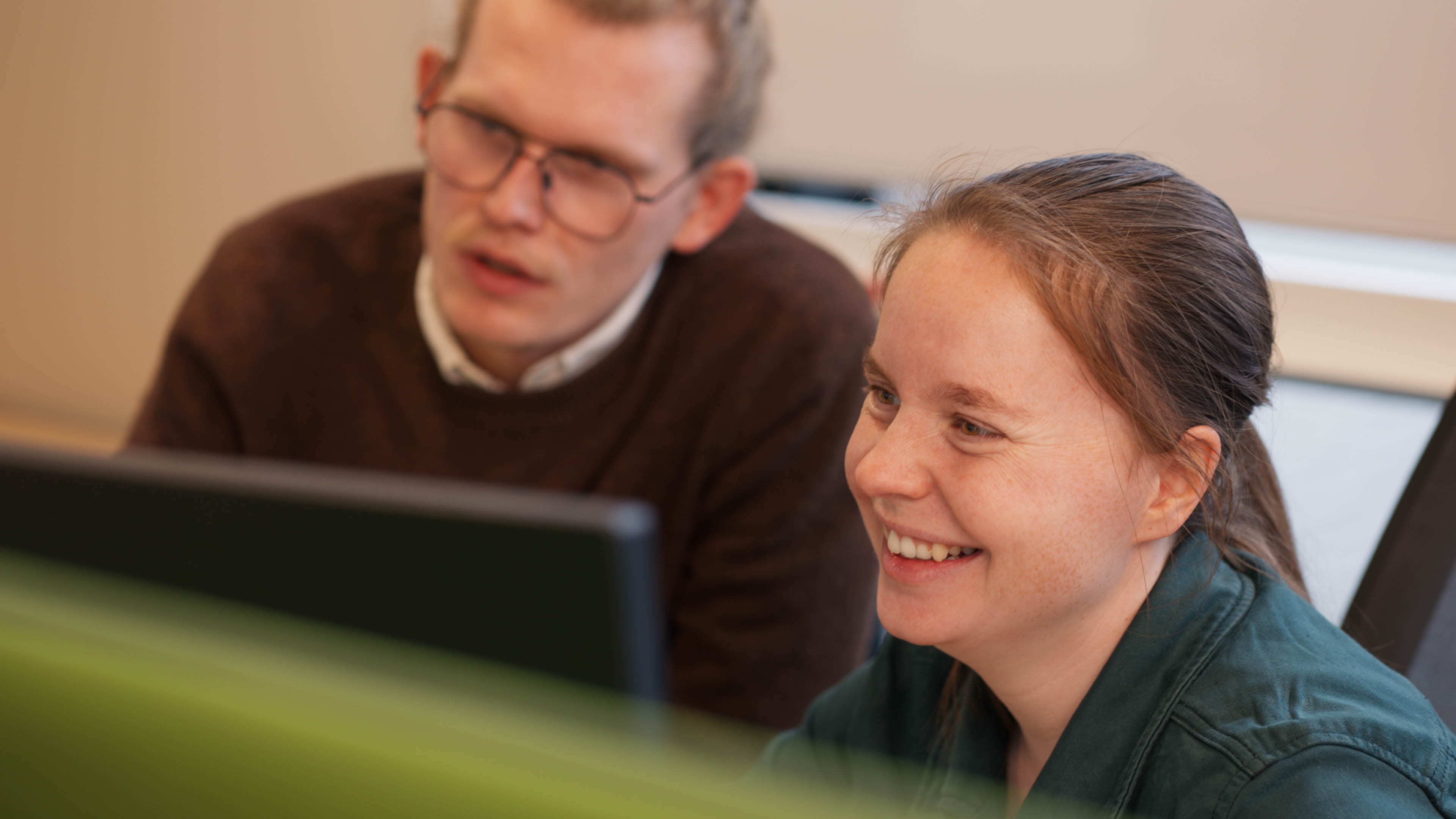 Twee collega’s werken samen en kijken naar een computerscherm, een vrouw lacht terwijl een man met bril naast haar zit.