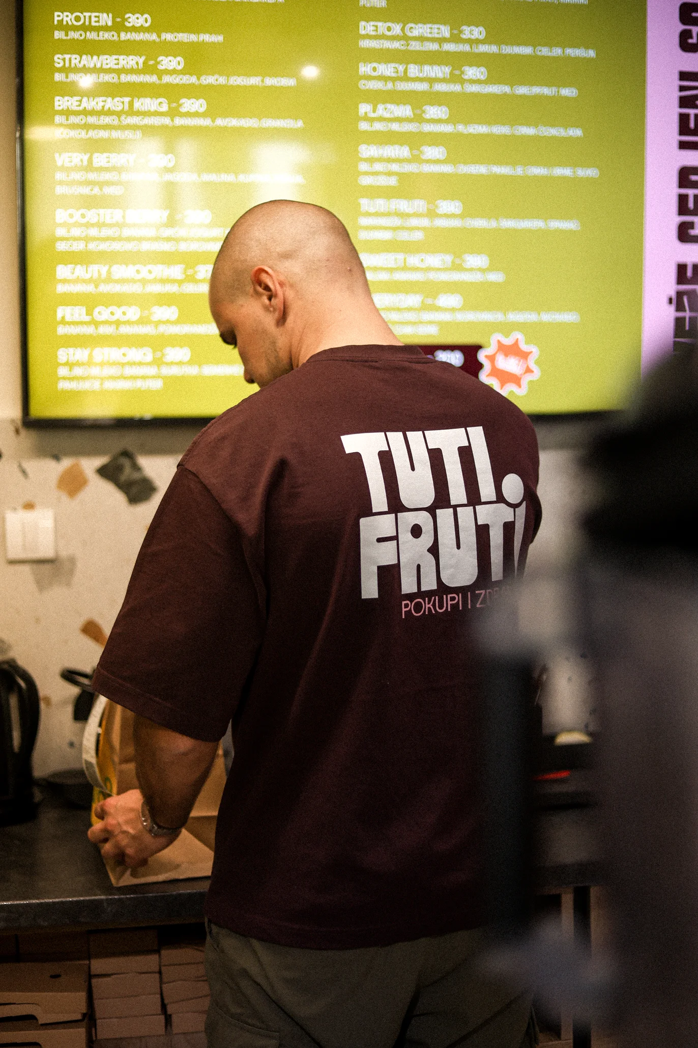 Man with shaved head wearing a maroon T-shirt with 'TUTI FRUTI' text, standing in front of a counter and a yellow menu board.