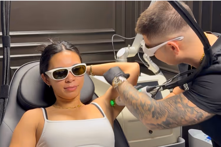 A woman wearing protective glasses receives a laser hair removal treatment on her underarm by a tattooed technician in a clinic.