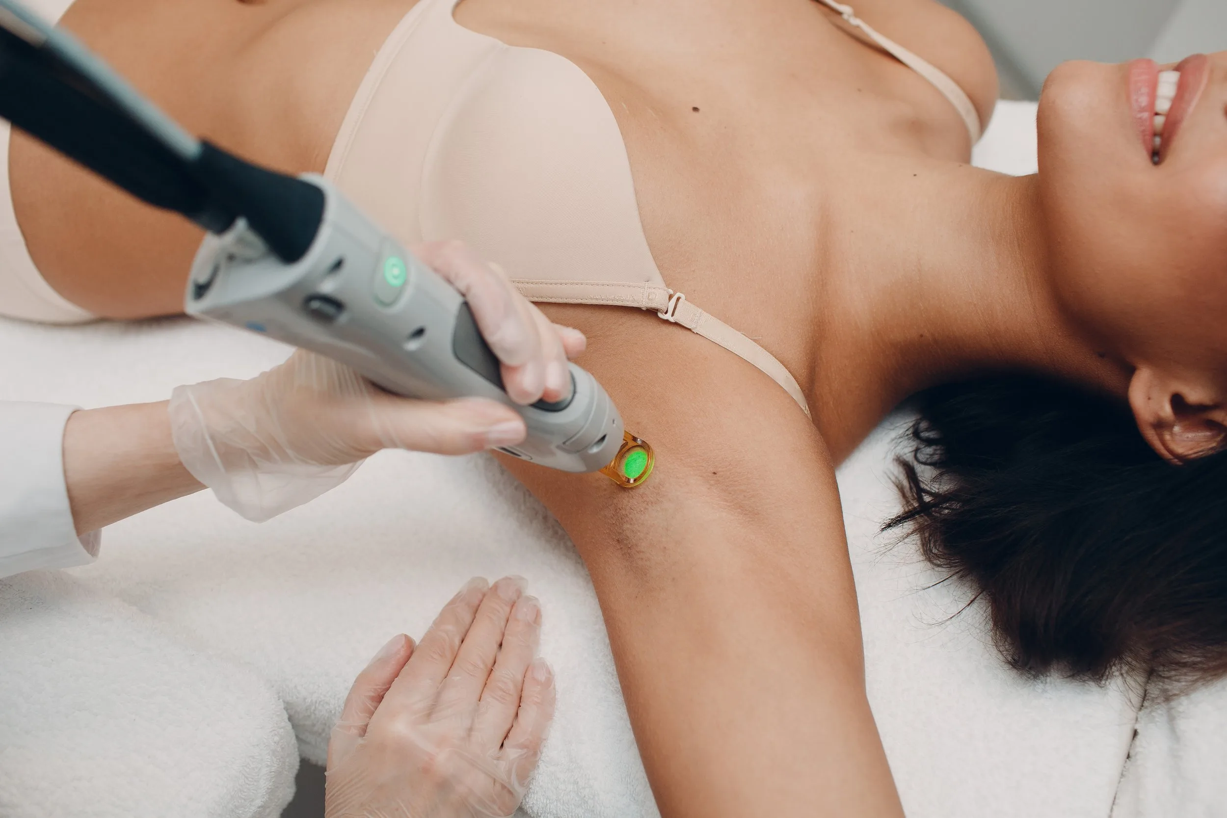 A woman wearing protective glasses receives a laser hair removal treatment on her underarm by a tattooed technician in a clinic.