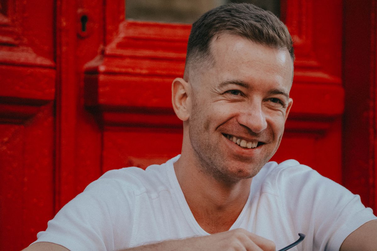 Portrait d'un homme souriant assis devant une porte d'entrée ancienne peinte en rouge