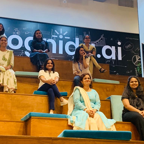 Group of seven women sitting on tiered wooden benches with teal cushions in front of a large wall sign displaying 'oid.ai' and illustrations of graphs and lightbulbs.
