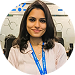 Smiling woman with dark hair wearing a white blouse and blue lanyard, standing indoors with a blurred background.