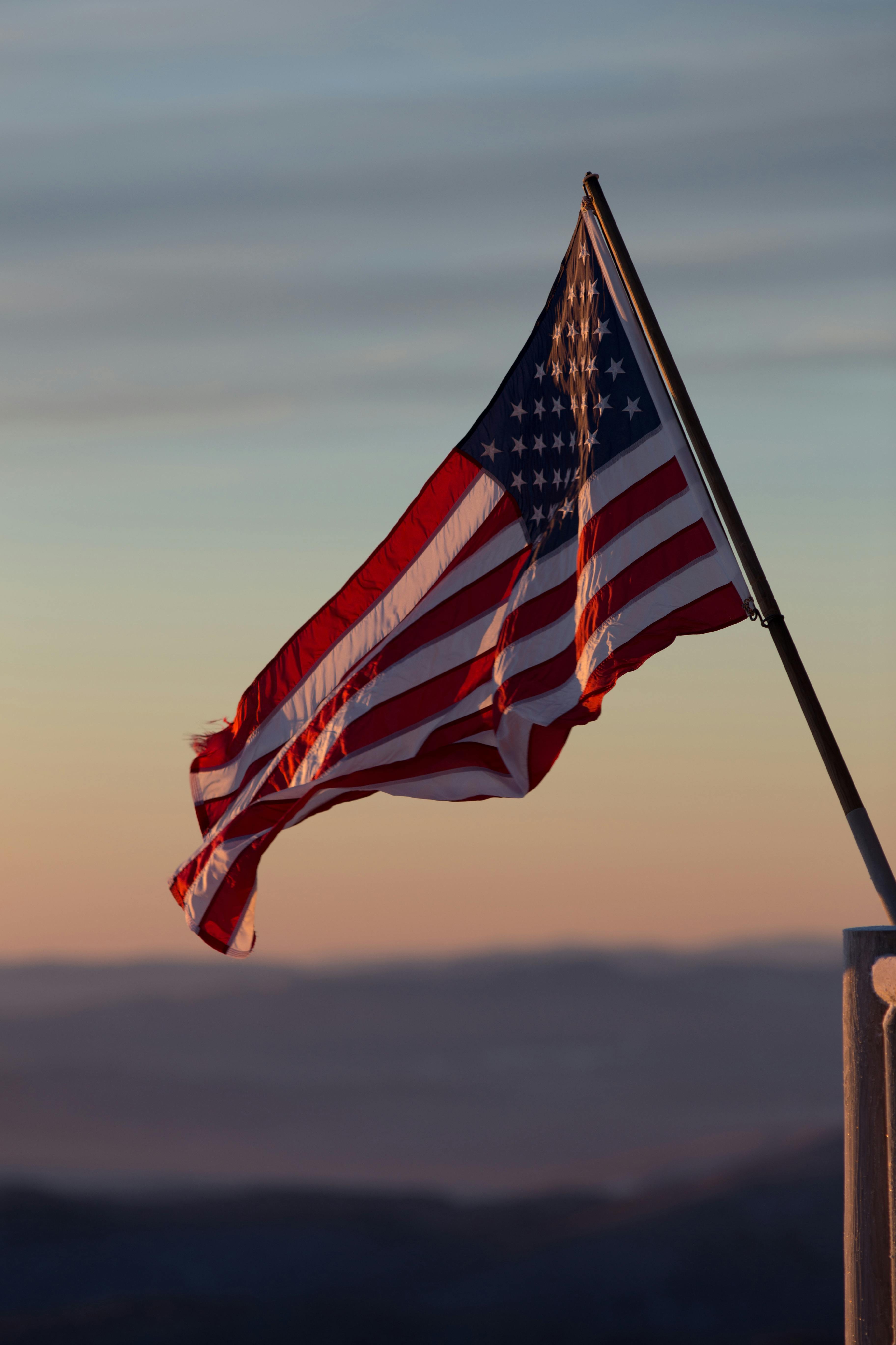 American flag waving on a pole against a sunset sky with distant hills.