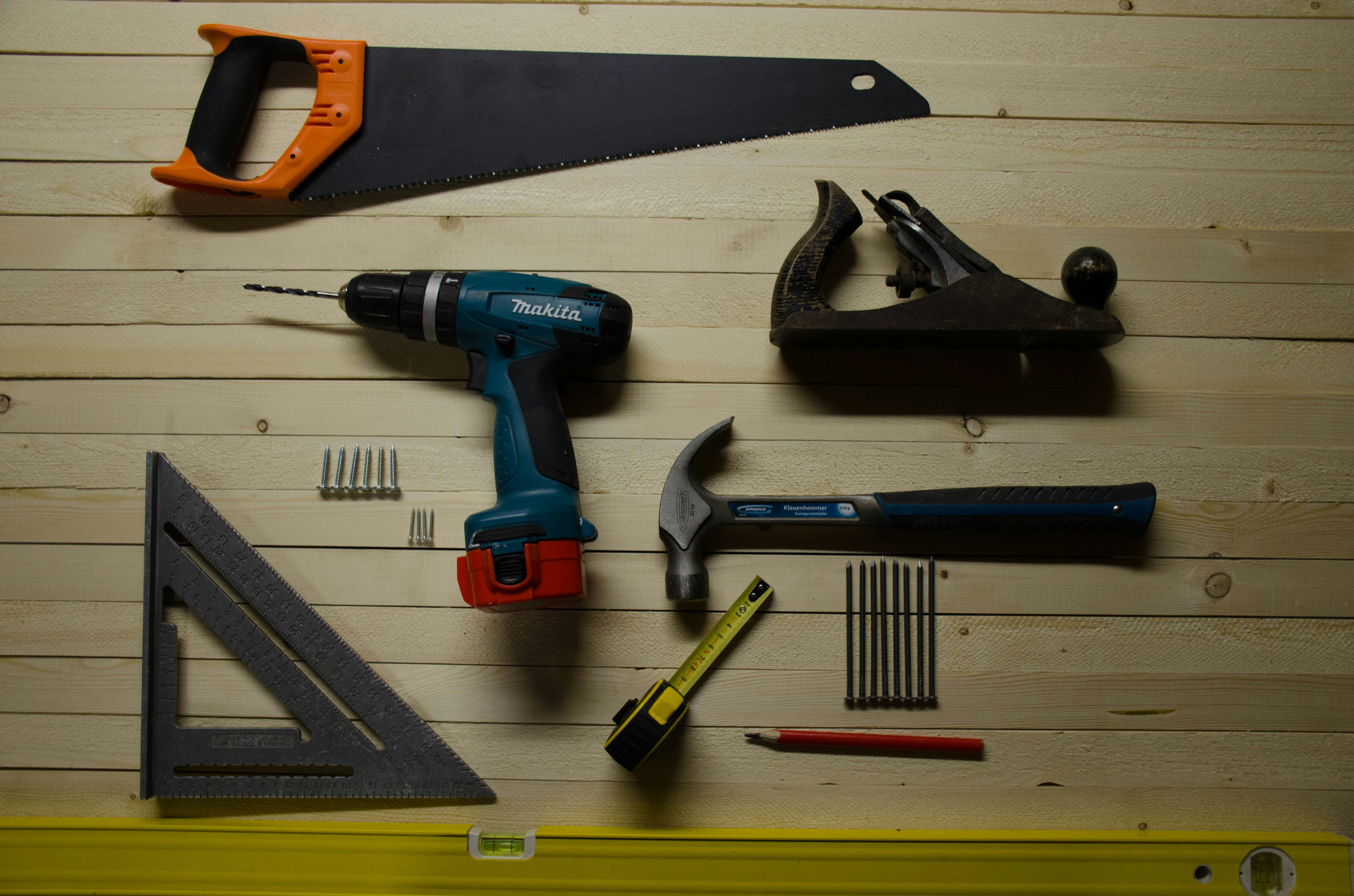 Various woodworking tools including saw, drill, hammer, tape measure, square, plane, nails, pencil on wood surface.