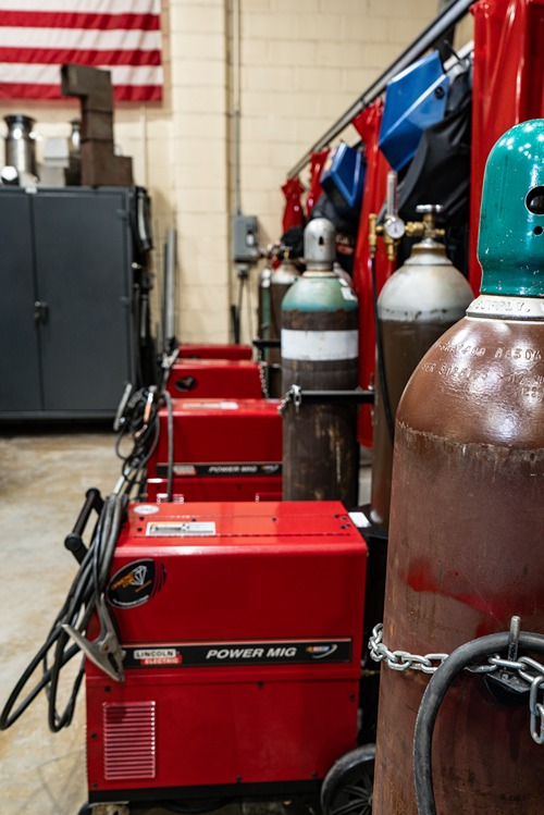 Row of red Lincoln Electric Power MIG welding machines and gas cylinders in a workshop.