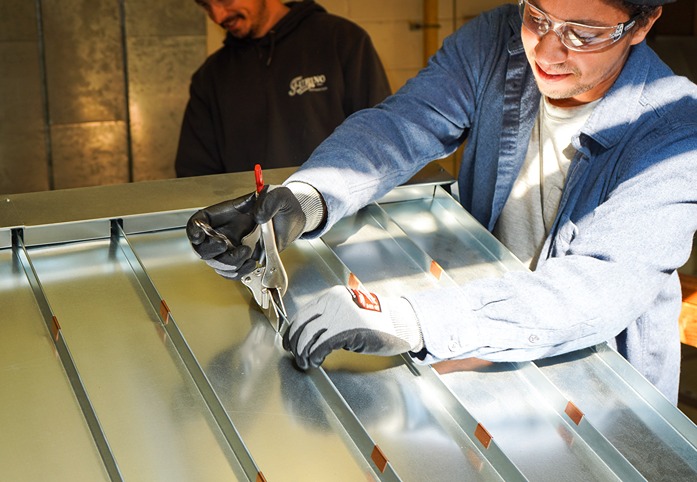 A man wearing safety glasses and gloves uses a clamp tool on a metal sheet while another man watches.