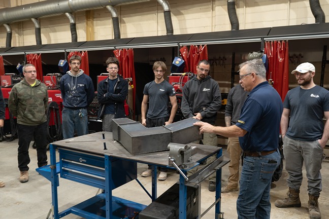 Instructor demonstrates metalwork bending to a group of students in a workshop with welding stations.