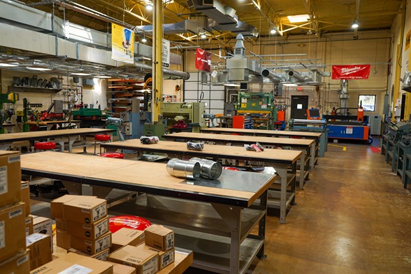 Industrial workshop with wooden tables, metal ducts, machinery, and stacked cardboard boxes.