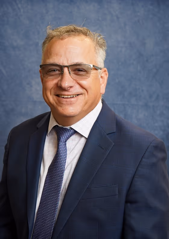 Smiling middle-aged man wearing glasses, blue suit, white shirt, and blue tie against a blue background.