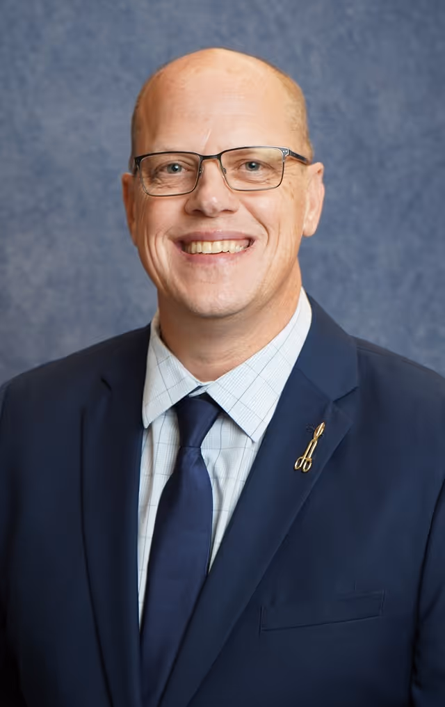 Smiling bald man with glasses, wearing a dark suit, checked shirt, and navy tie against blue background.