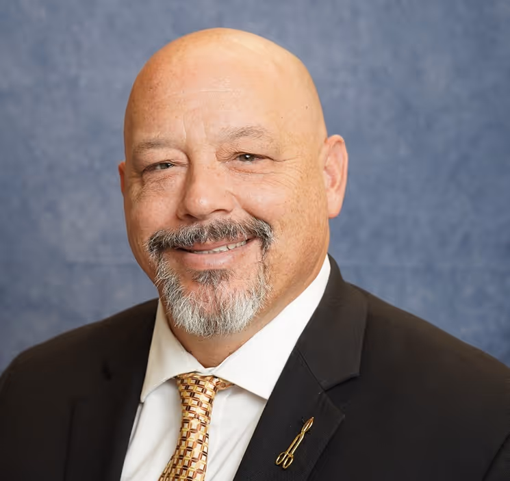 Smiling bald man with salt-and-pepper goatee wearing a suit, patterned tie, and a pen-shaped lapel pin.