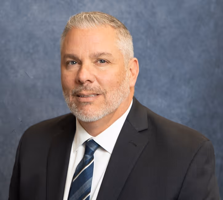 Middle-aged man with gray hair and beard wearing a dark suit and striped tie against a blue background.