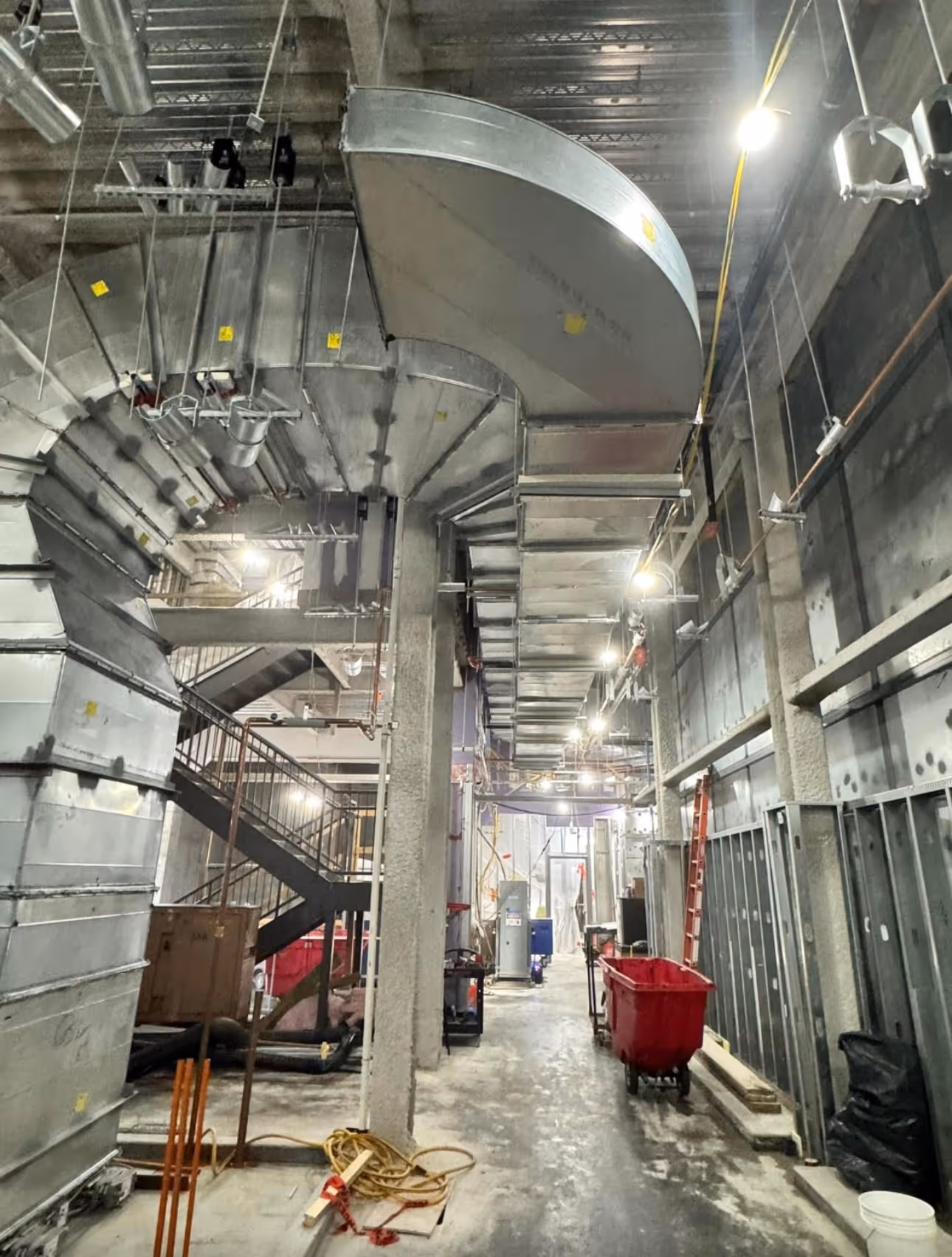 Interior view of a construction site with exposed ductwork, stairs, red bins, and building materials.